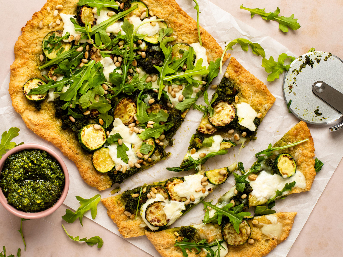 This is a horizontal overhead image of a rectangular shaped crust dolloped with pesto and shredded cheese, browned zucchini coins, burrata cheese, arugula and toasted pine nuts. The flatbread sits on a white piece of parchment paper on a light pink surface and a few triangular slides are cut from the crust. A small pink bowl filled with pesto is to the bottom left of the image and a pizza cutter is at the top right corner.