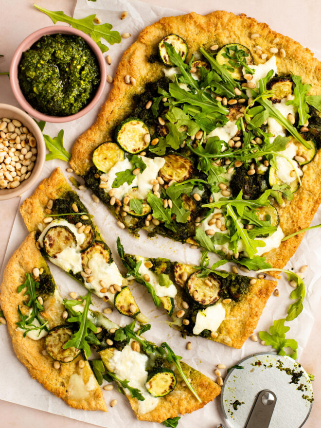 This is a vertical overhead image of a rectangular shaped crust dolloped with pesto and shredded cheese, browned zucchini coins, burrata cheese, arugula and toasted pine nuts. The flatbread sits on a white piece of parchment paper on a light pink surface and a few triangular slides are cut from the crust. A small pink bowl filled with pesto is to the top left of the image and another small bowl filled with toasted pine nuts is next to it. A pizza cutter is at the bottom right corner.