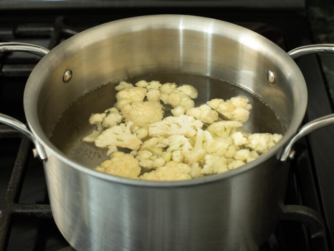 This is a horizontal image looking into a silver cooking pot filled with water and cauliflower florets floating in it. The pot is sitting on a stovetop.