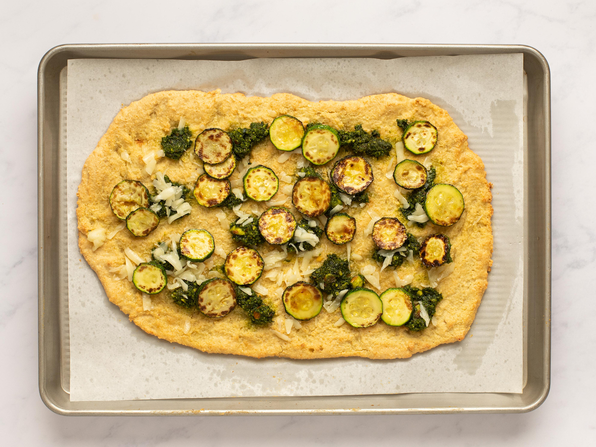 This is an overhead horizontal image of a baking sheet with white parchment paper on a white marble surface. On the parchment paper is a rectangular shaped crust dolloped with pesto and shredded cheese and browned zucchini coins.