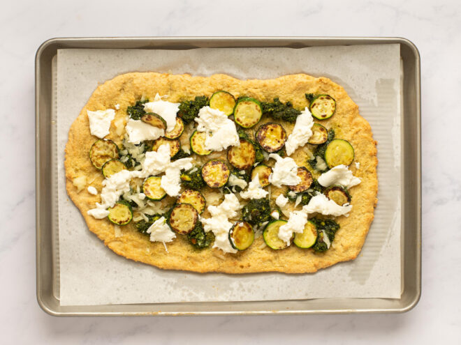 This is an overhead horizontal image of a baking sheet with white parchment paper on a white marble surface. On the parchment paper is a rectangular shaped crust dolloped with pesto and shredded cheese, browned zucchini coins and burrata cheese.