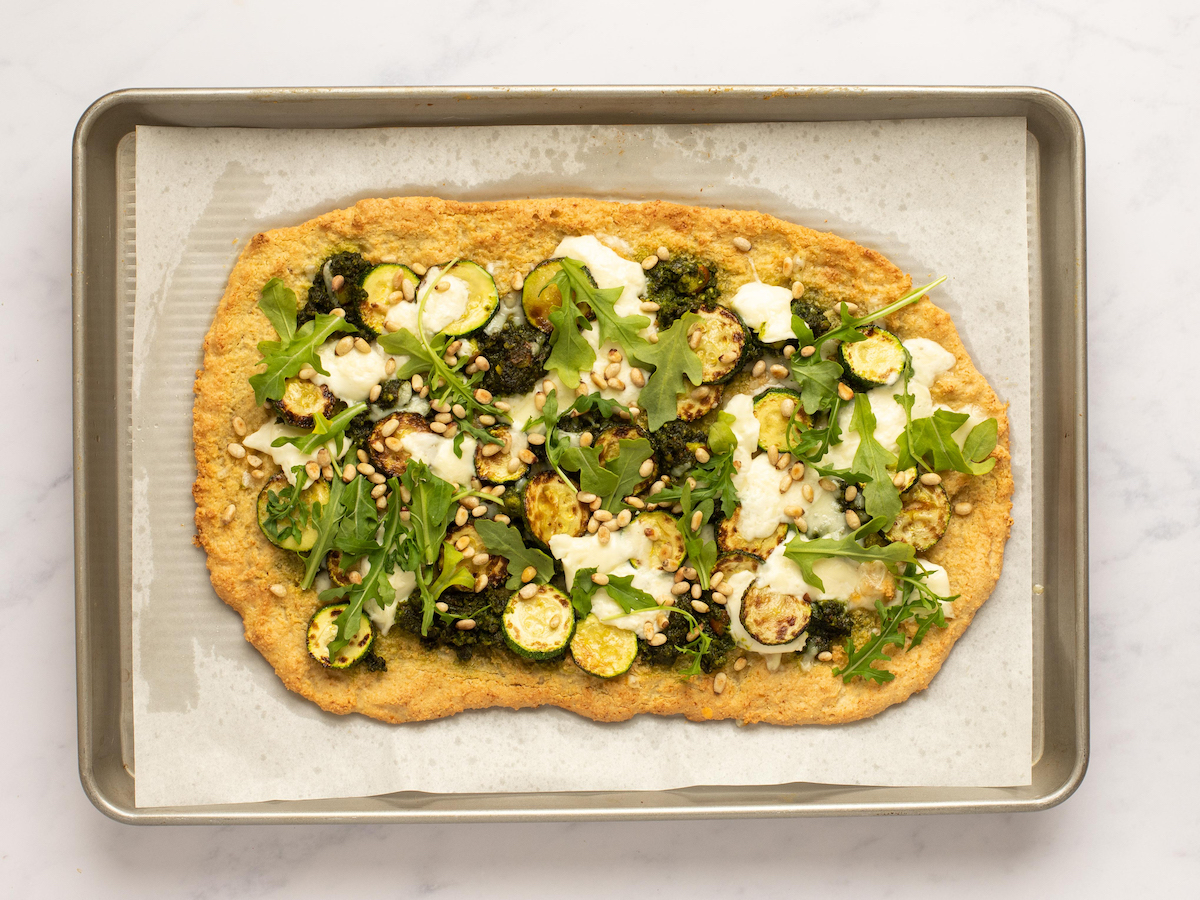 This is an overhead horizontal image of a baking sheet with white parchment paper on a white marble surface. On the parchment paper is a rectangular shaped crust dolloped with pesto and shredded cheese, browned zucchini coins, burrata cheese, arugula and toasted pine nuts.
