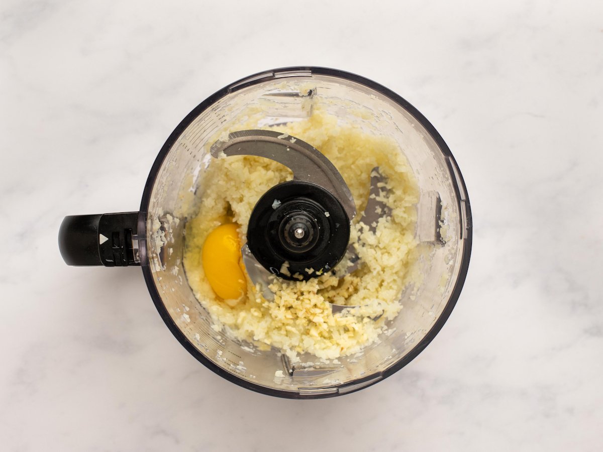This is an overhead horizontal image of a food processor with cauliflower rice chopped up in it and an egg. The food processor sits on a white marble surface.