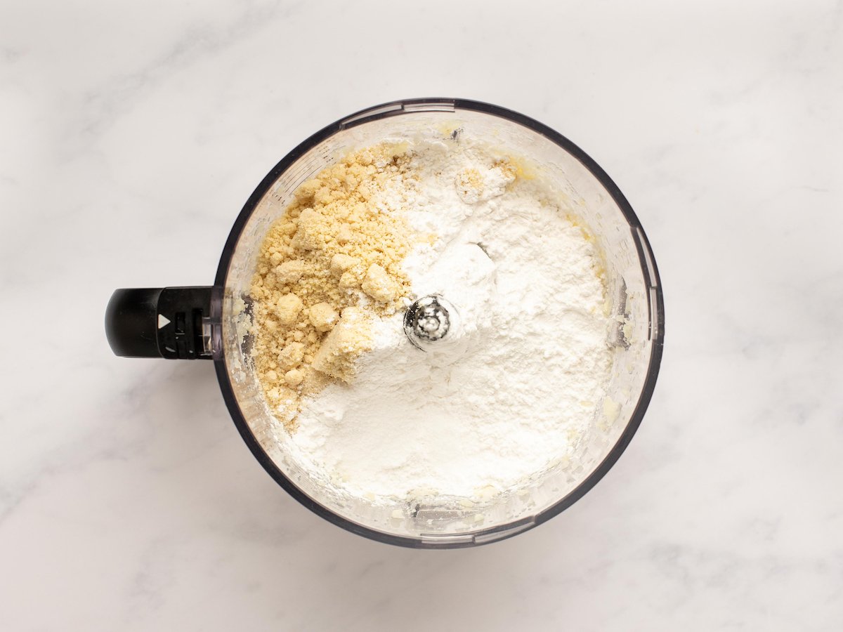 This is an overhead horizontal image of a food processor with blanched almond flour and arrowroot flour in it. The food processor sits on a white marble surface.