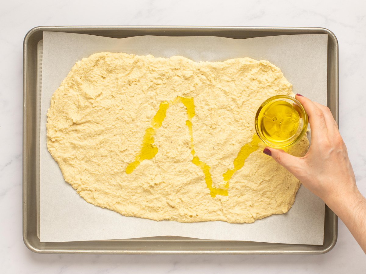 This is an overhead horizontal image of a baking sheet with white parchment paper. On the parchment paper is uncooked dough spread out in a rectangular shape. A hand is coming from the right side of the image holding a small glass bowl of olive oil and drizzling it onto the crust.