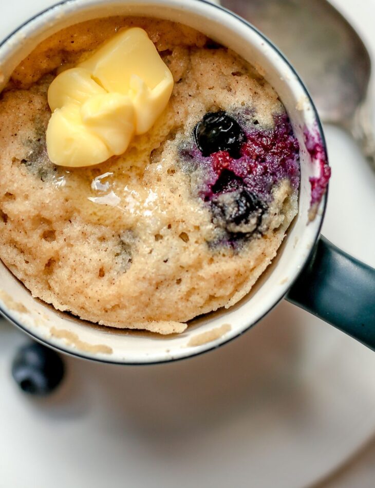 This is an overhead horizontal image of a tall grey mug with a white inside sitting on a small white plate with a silver spoon laying beside it. The plate sits on a white surface. In the mug is a blueberry cake/muffin topped with a pad of butter that is slightly melted.