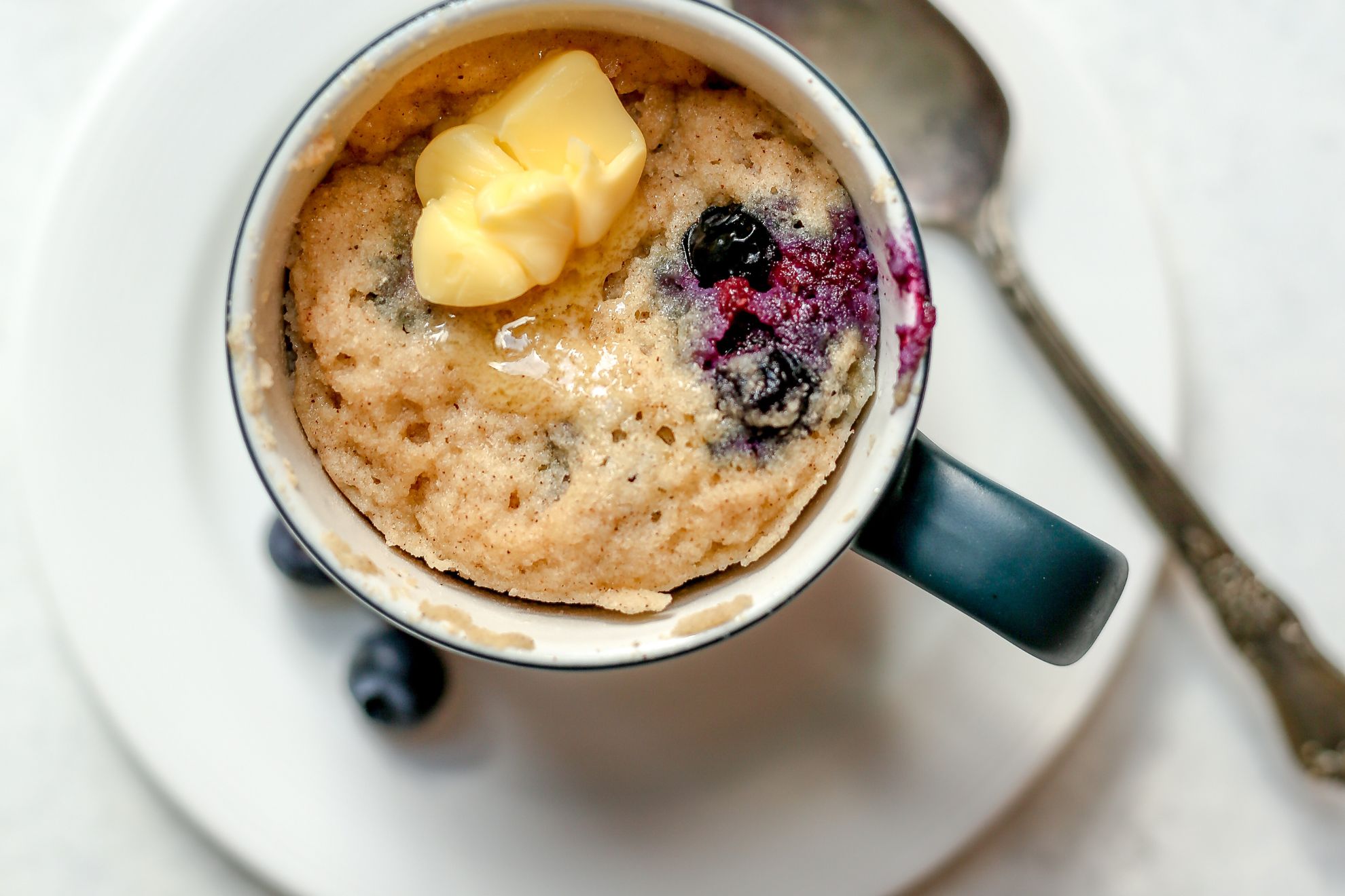 This is an overhead horizontal image of a tall grey mug with a white inside sitting on a small white plate with a silver spoon laying beside it. The plate sits on a white surface. In the mug is a blueberry cake/muffin topped with a pad of butter that is slightly melted.