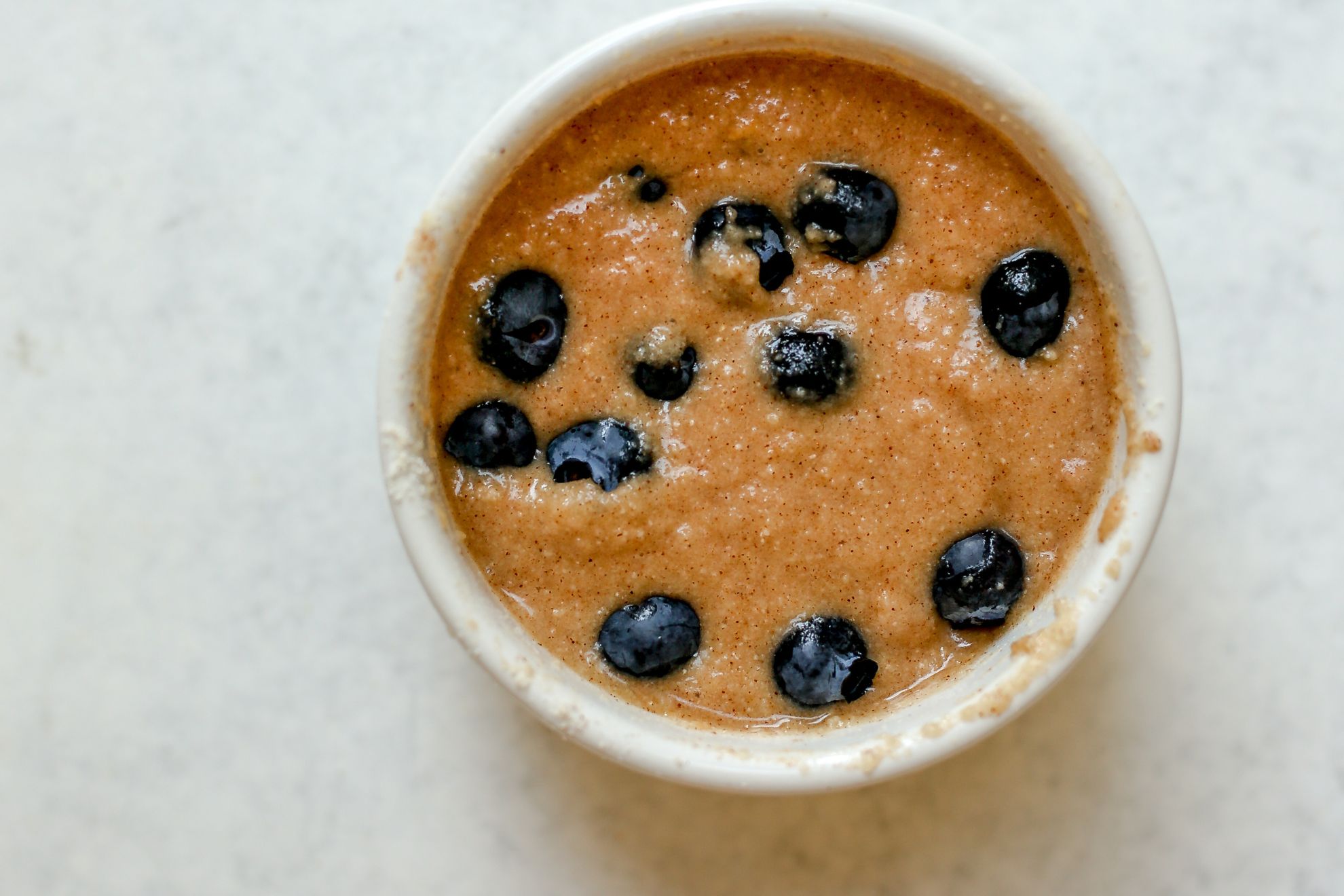 This is an overhead horizontal image of a small white bowl with raw muffin batter and blueberries on top of it. The bowl sits on a white surface.