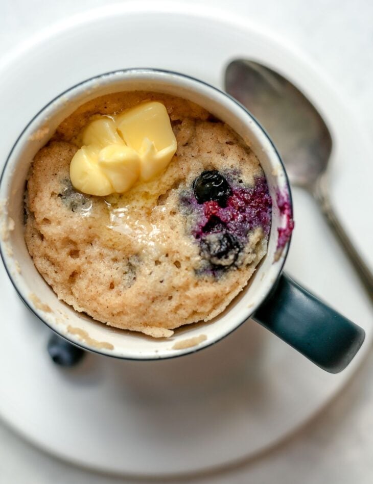This is an overhead vertical image of a tall grey mug with a white inside sitting on a small white plate with a silver spoon laying beside it. The plate sits on a white surface. In the mug is a blueberry cake/muffin topped with a pad of butter that is slightly melted.