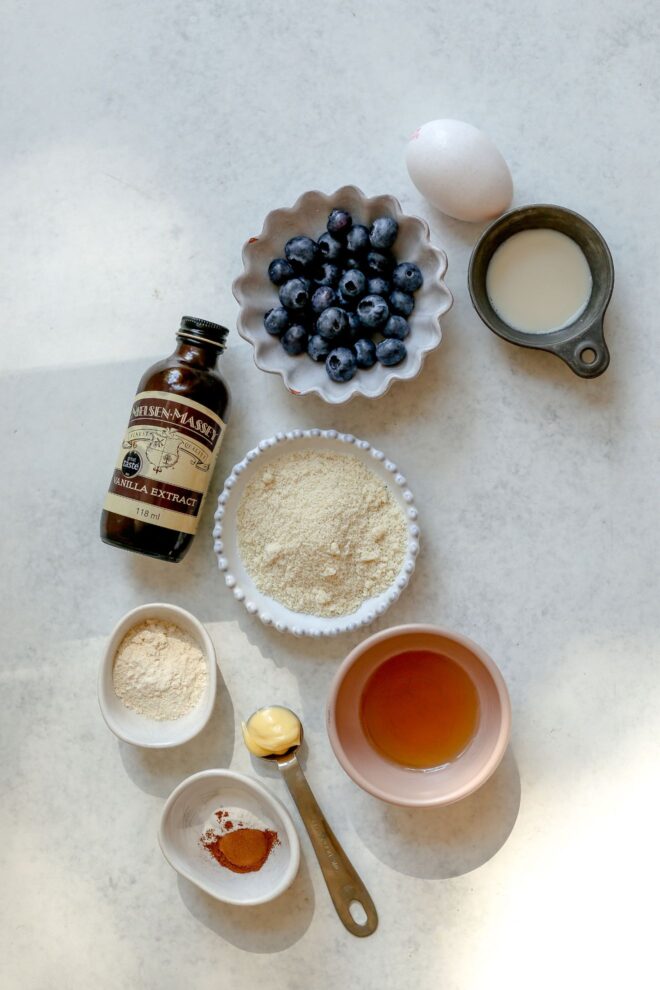 This is an overhead vertical image of an egg, a measuring cup with almond milk, a small bowl of blueberries, a vanilla extract bottle, a small bowl of almond flour, a small bowl of coconut flour, a small bowl of honey, a teaspoon of butter, and a small bowl with salt and cinnamon.