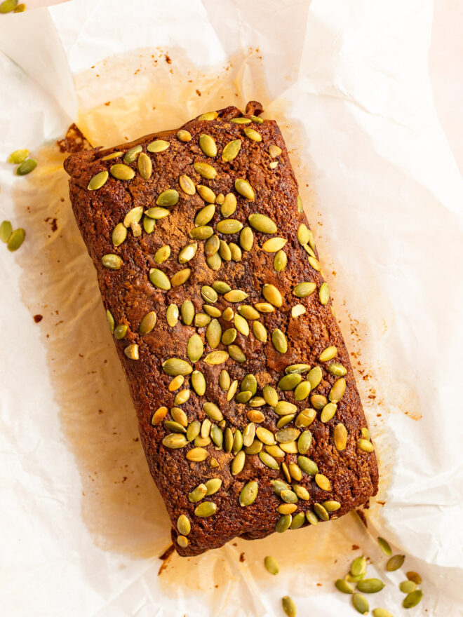 This is an overhead vertical image of a pumpkin bread topped with green pumpkin seeds sitting on a white piece of parchment paper. The bread is vertical but tilted slightly to the left top corner of the image.