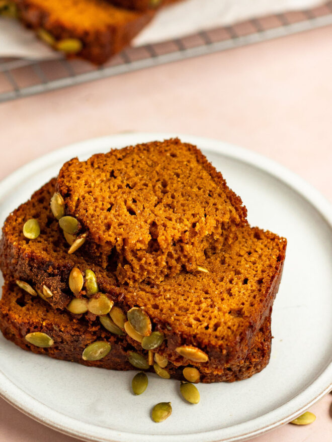 This is a vertical image looking at a small white plate from the side. On the plate is a stack of three slices of pumpkin bread, the top slice is broken in half. Green pumpkin seeds are on the top of each slice of bread. The plate sits on a light pink surface with a cooling rack with the pumpkin bread on it blurred in the background.