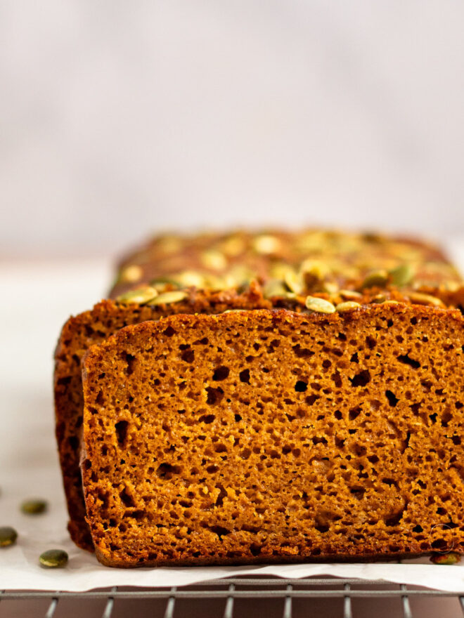This is a horizontal image looking at pumpkin bread loaf sitting on a piece of white parchment paper on a cooling rack. The pumpkin bread has green pumpkin seeds on top and a few have fallen off and are scattered around the base of the bread. The bread is cut into revealing the fluffy texture. The cooling rack sits on a light colored surface with a white background.