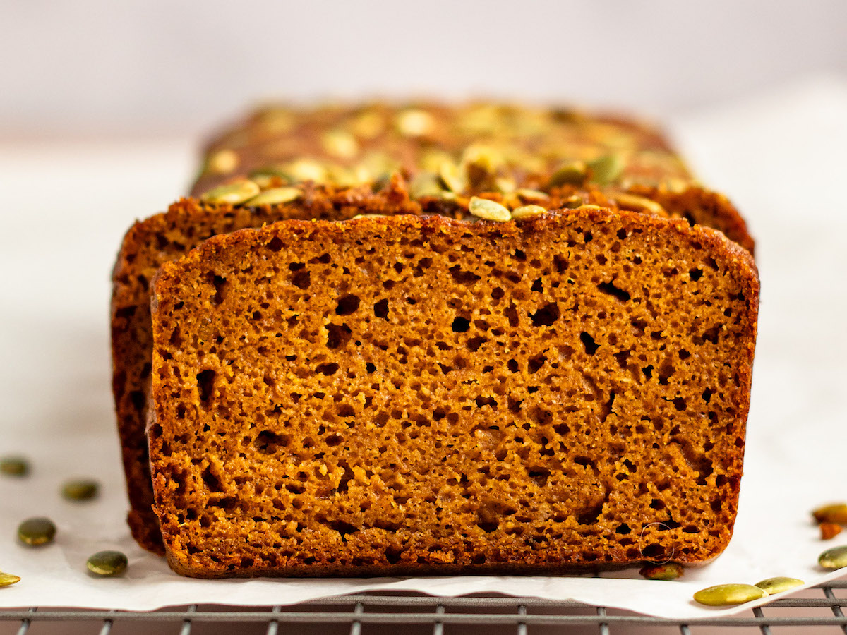 This is a horizontal image looking at pumpkin bread loaf sitting on a piece of white parchment paper on a cooling rack. The pumpkin bread has green pumpkin seeds on top and a few have fallen off and are scattered around the base of the bread. The bread is cut into revealing the fluffy texture. The cooling rack sits on a light colored surface with a white background.