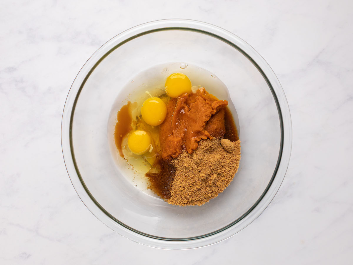 This is an overhead horizontal image of a glass bowl on a white marble surface. In the bowl are eggs, pumpkin puree, coconut sugar, and vanilla extract.