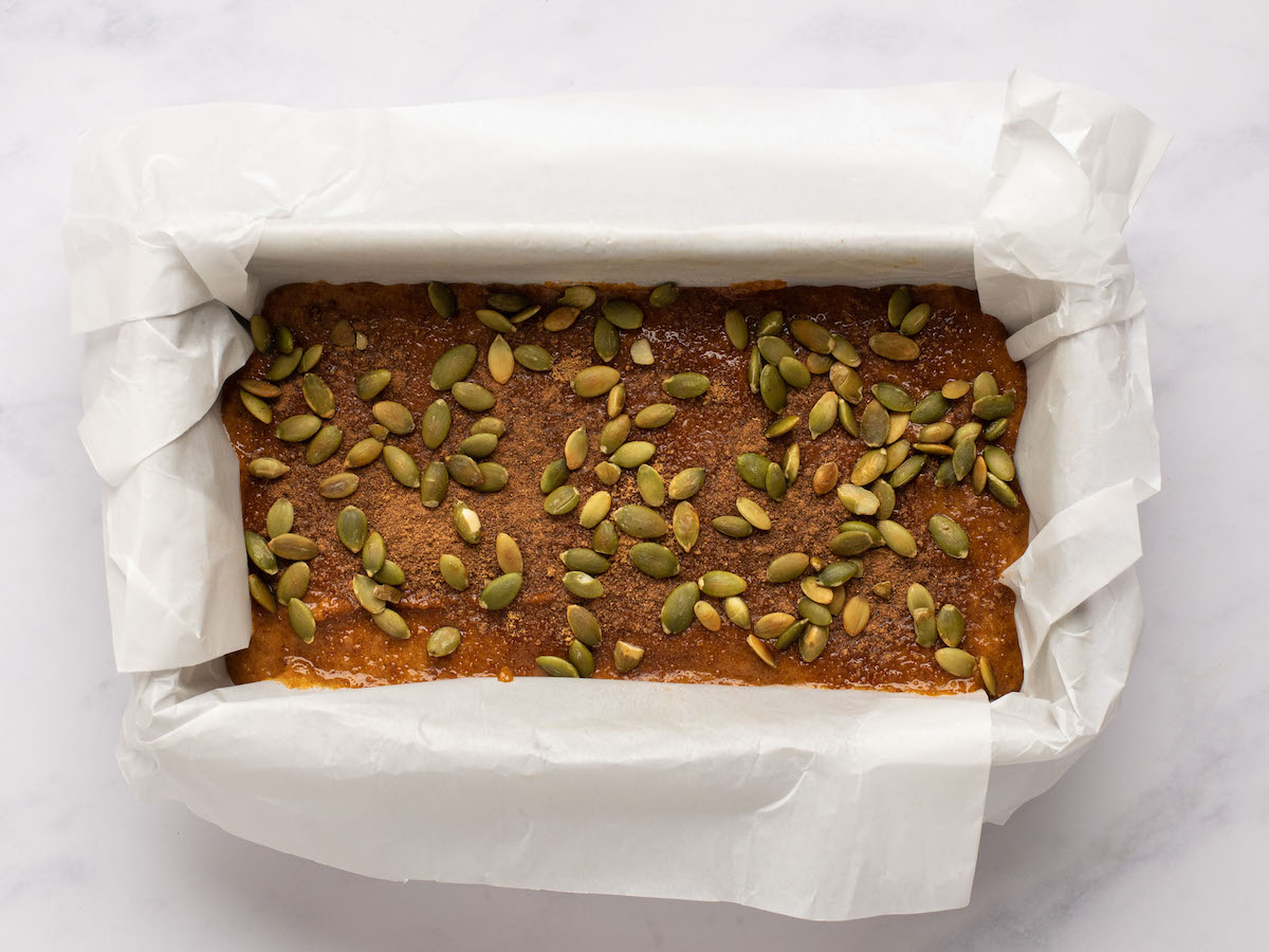 This is an overhead horizontal image of a bread pan lined with parchment paper. In the pan is a raw bread batter topped with pumpkin seeds. The bread pan sits on a white marble surface.