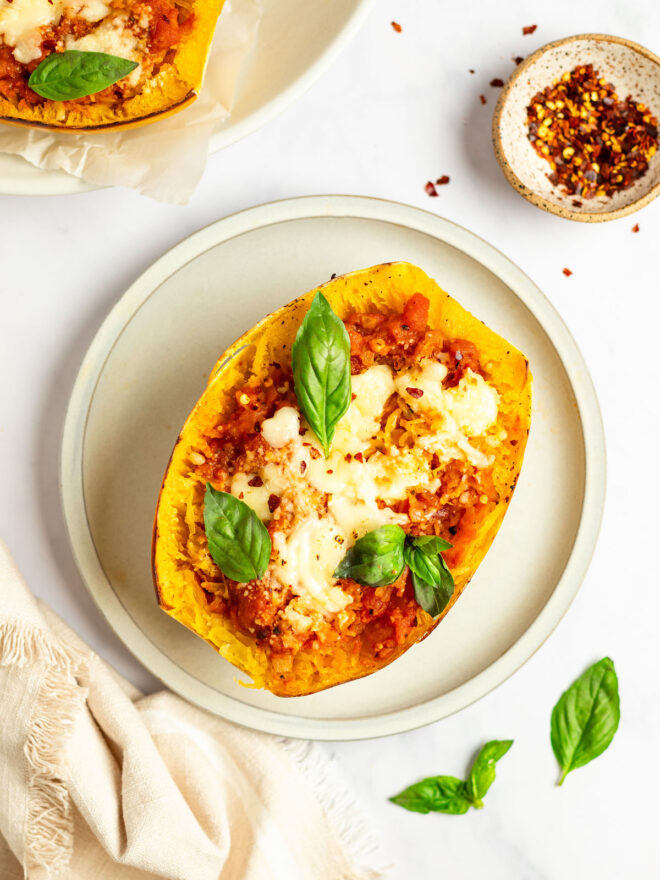 This is an overhead vertical image of half of a baked spaghetti squash with an orange-red marinara sauce in it, melted cheese and a couple basil leaves. The squash sits on a white plate on a white surface with a small bowl of red pepper flakes to the top right corner, another squash peeking in from the left top corner, a small beige tea towel in the bottom left corner and basil leaves in the right bottom corner.