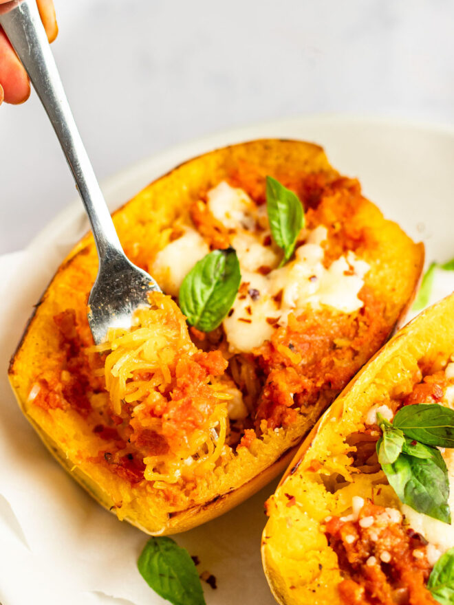 This is a vertical image looking at a roasted spaghetti squash from the side. A fork is pulling up some strands from the spaghetti squash half. The spaghetti squash is topped with an orange-red sauce, melted cheese, basil leaves and red pepper flakes. The spaghetti squash half sits on a white surface with another half peeking in from the bottom right corner.