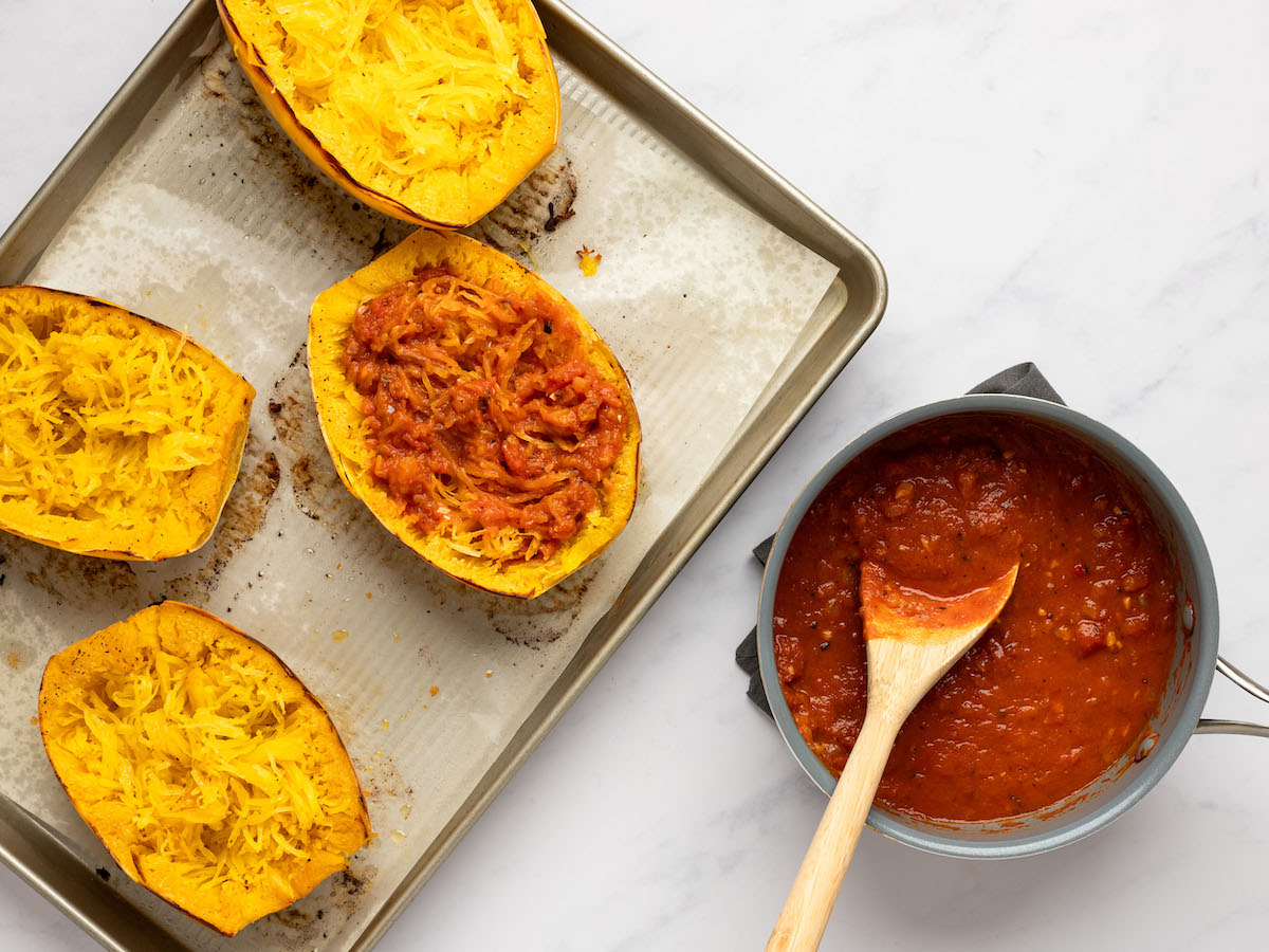 This is an overhead horizontal image of four halves of a spaghetti squash sitting on a piece of parchment paper on a rimmed baking sheet. The baking sheet sits on a white marble surface and is angled to the left side of the image. One spaghetti squash half has red pasta sauce in it. To the bottom right of the image is a small saucepan on a dark grey tea towel. In the pan is a red marinara sauce with a wooden spoon dipping into the sauce. The wooden handle is leaning against the side of the pan and pointing to the bottom center of the image.