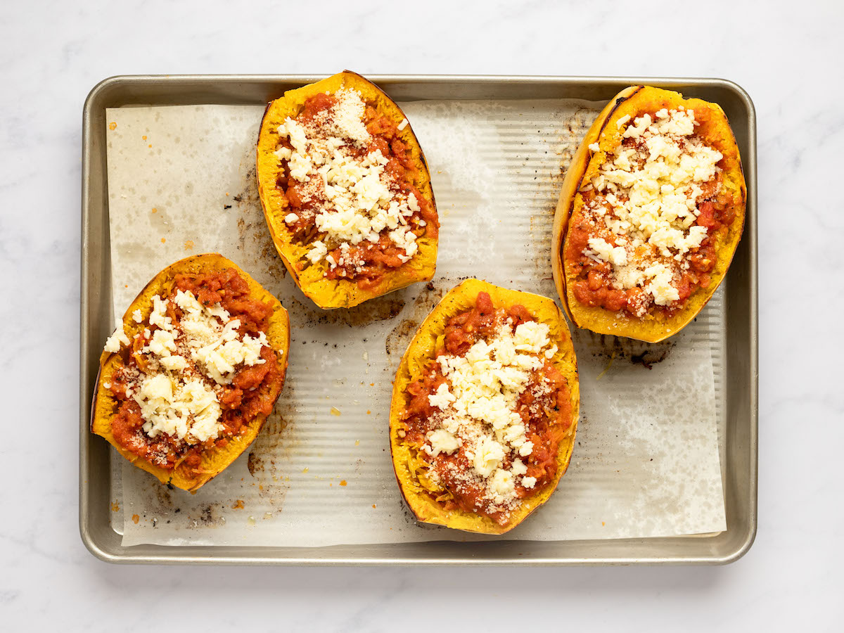 This is an overhead horizontal image of four halves of a spaghetti squash sitting on a piece of parchment paper on a rimmed baking sheet. The baking sheet sits on a white marble surface. The spaghetti squash halves are topped with a marinara sauce and crumbles of cheese.