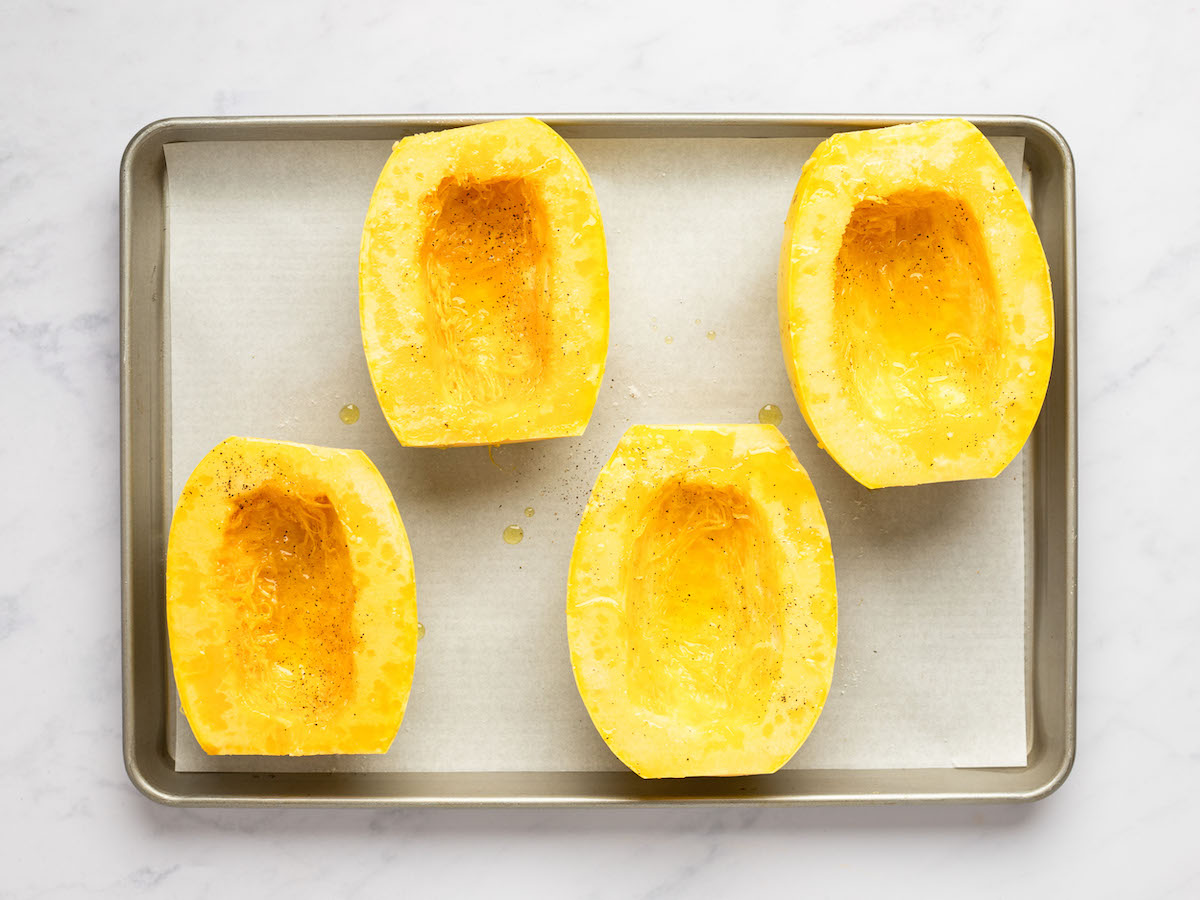 This is an overhead horizontal image of a rimmed baking sheet lined with parchment paper. On the parchment paper are four halves of spaghetti squash facing up with oil, salt and pepper on them. The baking sheet is on a white marble surface.