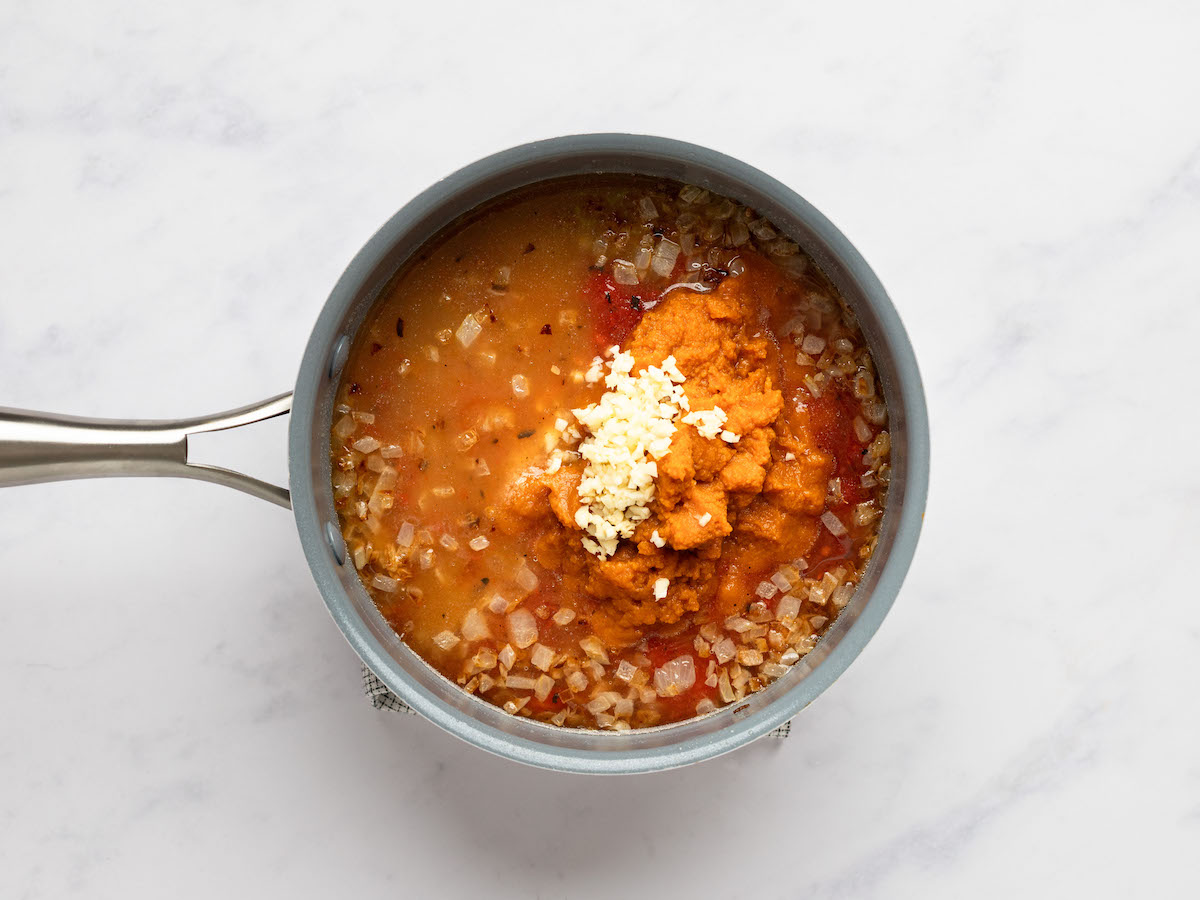 This is an overhead horizontal image of a saucepan with onions, brother, tomato sauce, pumpkin puree and garlic in it. The pot sits on a white marble surface with a small check tea towel peeking out from underneath.