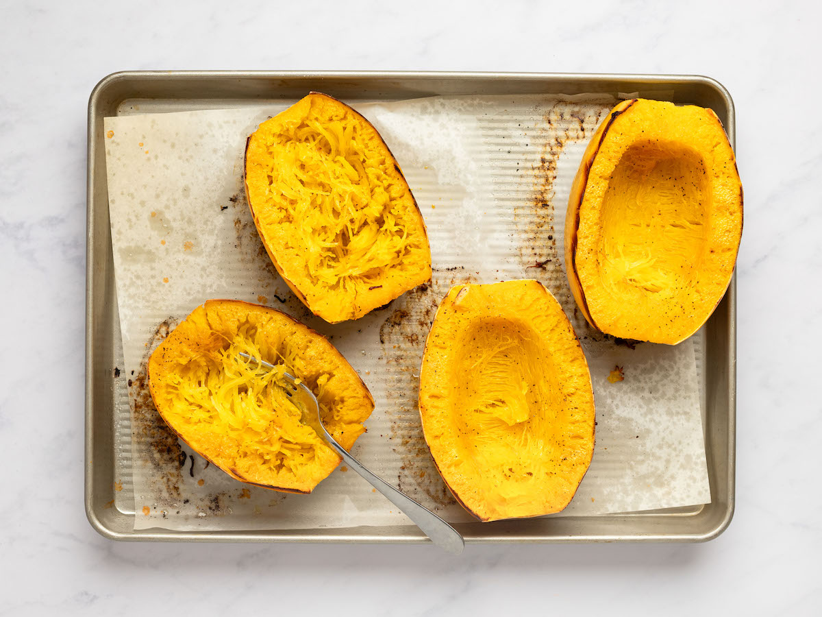 This is an overhead horizontal image of four halves of a spaghetti squash sitting on a piece of parchment paper on a rimmed baking sheet. The baking sheet sits on a white marble surface. The spaghetti squash halves are facing up. Two halves have the spaghetti strands pulled away from the sides of the shell with a fork in one of them.
