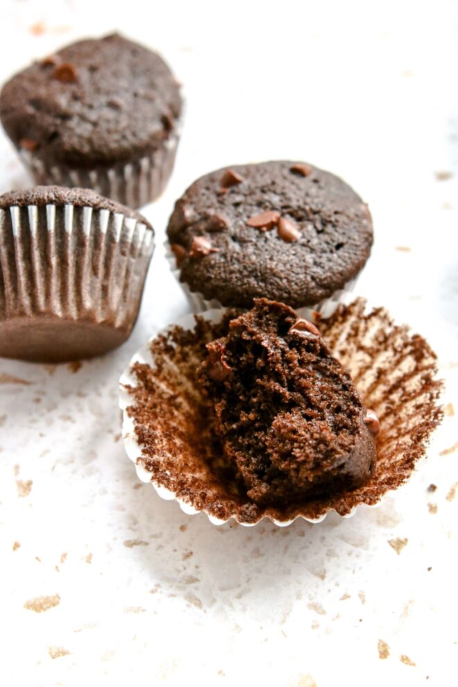This is a vertical image looking at a chocolate mini muffin from an angle. The chocolate muffin has a bite taken out of it and is leaning on its side on top of its white parchment liner. More muffins are behind the muffin. All the muffins sit on a white terrazzo surface.