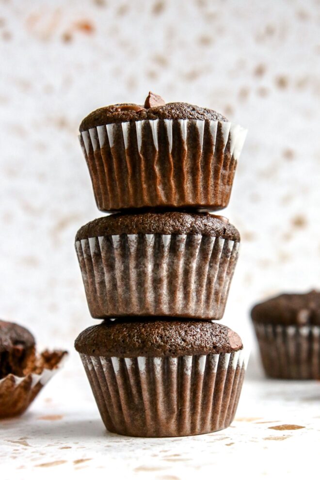 This is a vertical image looking at a stack of three chocolate muffins each with white parchment paper liners. The muffins sit on a white terrazzo surface with more muffins blurred to either side in the background.