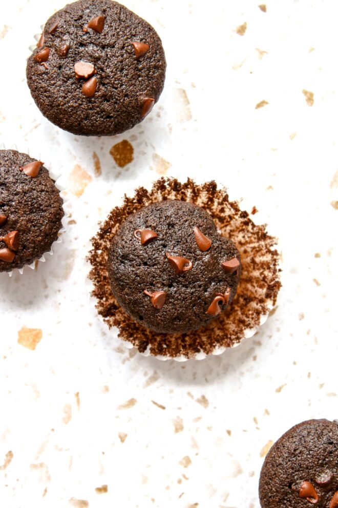 This is an overhead vertical image of centering on one mini muffin. The white parchment liner is pulled away from the muffin. More mini muffins are to the top left corner and bottom right corner of the image. The muffins sit on a white terrazzo surface.