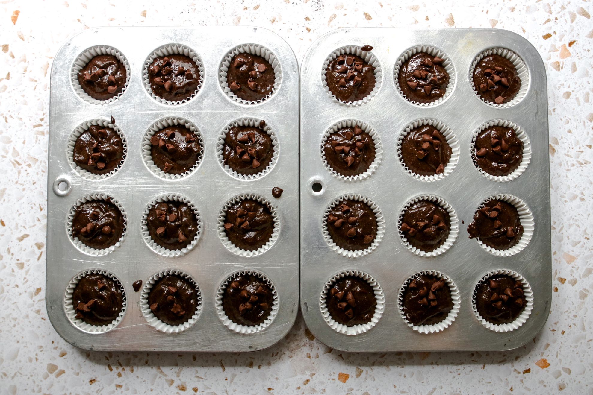 This is an overhead horizontal image of two silver mini muffin tins on a white terrazzo surface. In each muffin tin cup are white parchment liners and a chocolate batter, each topped with mini chocolate chips.