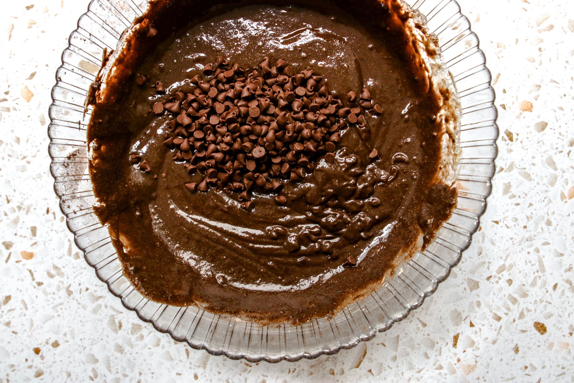 This is an overhead horizontal image of a large glass bowl with scalloped edges on a white terrazzo surface. In the bowl is a raw chocolate batter topped with a mound of mini chocolate chips.