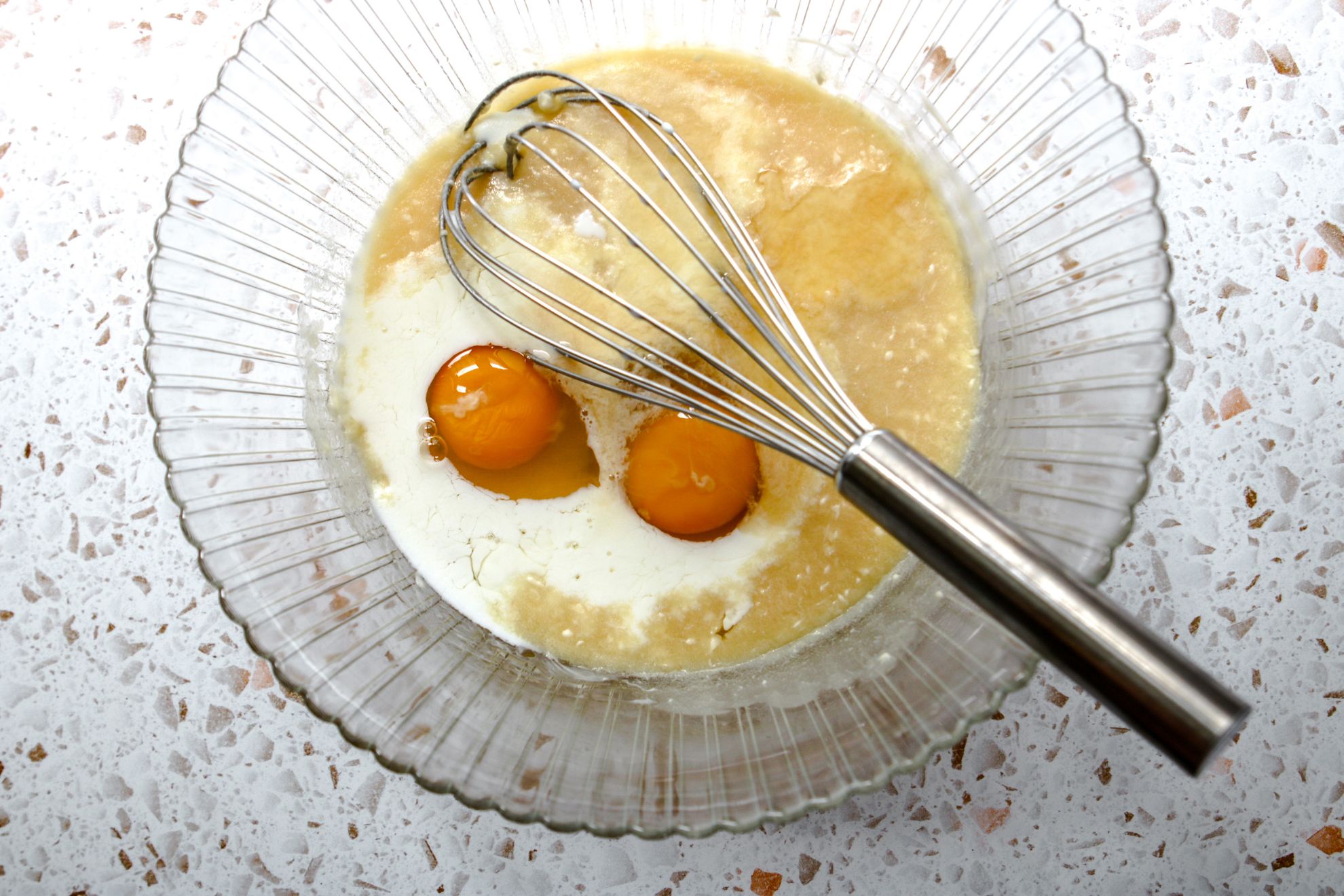 This is an overhead horizontal image of a large glass bowl with scalloped edges on a white terrazzo surface. In the bowl or wet ingredients including milk and eggs. A silver whisk is in the bowl with the silver handle leaning against the side of the bowl pointing to the bottom right corner.