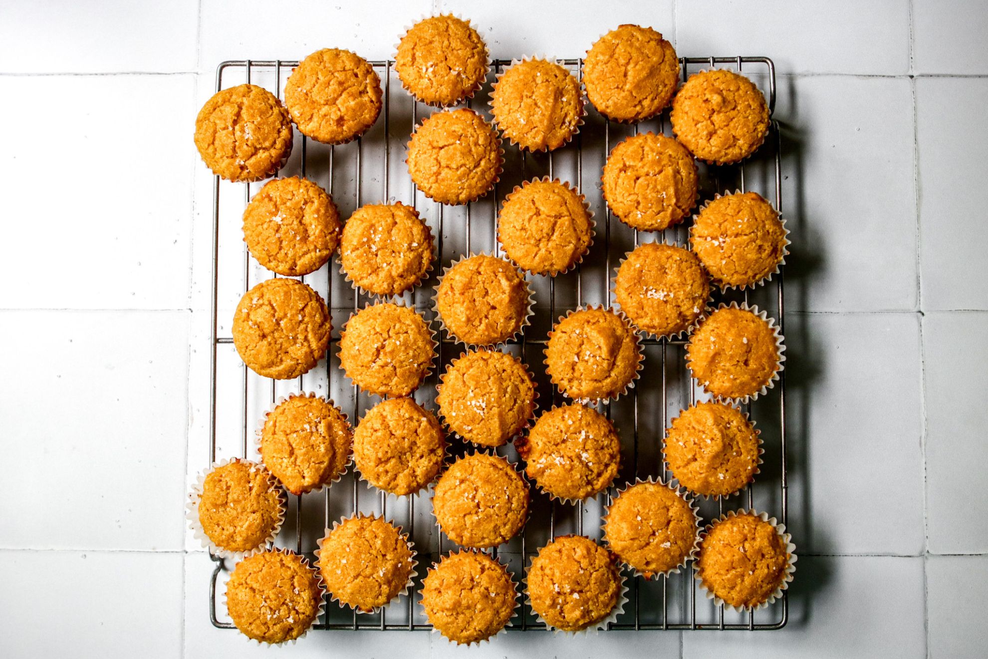 This is an overhead horizontal image of a silver cooling rack with baked mini cornbread muffins on it. The mini muffins are topped with flakey salt. The cooling rack sits on a white square tiled surface.