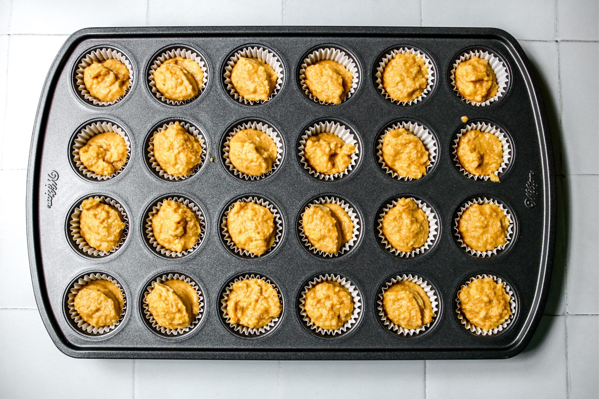 This is an overhead horizontal image of a 24-cup mini muffin tin with white parchment paper muffin liners and raw cornbread batter in each cup. The muffin tin sits on a white square tiled surface.