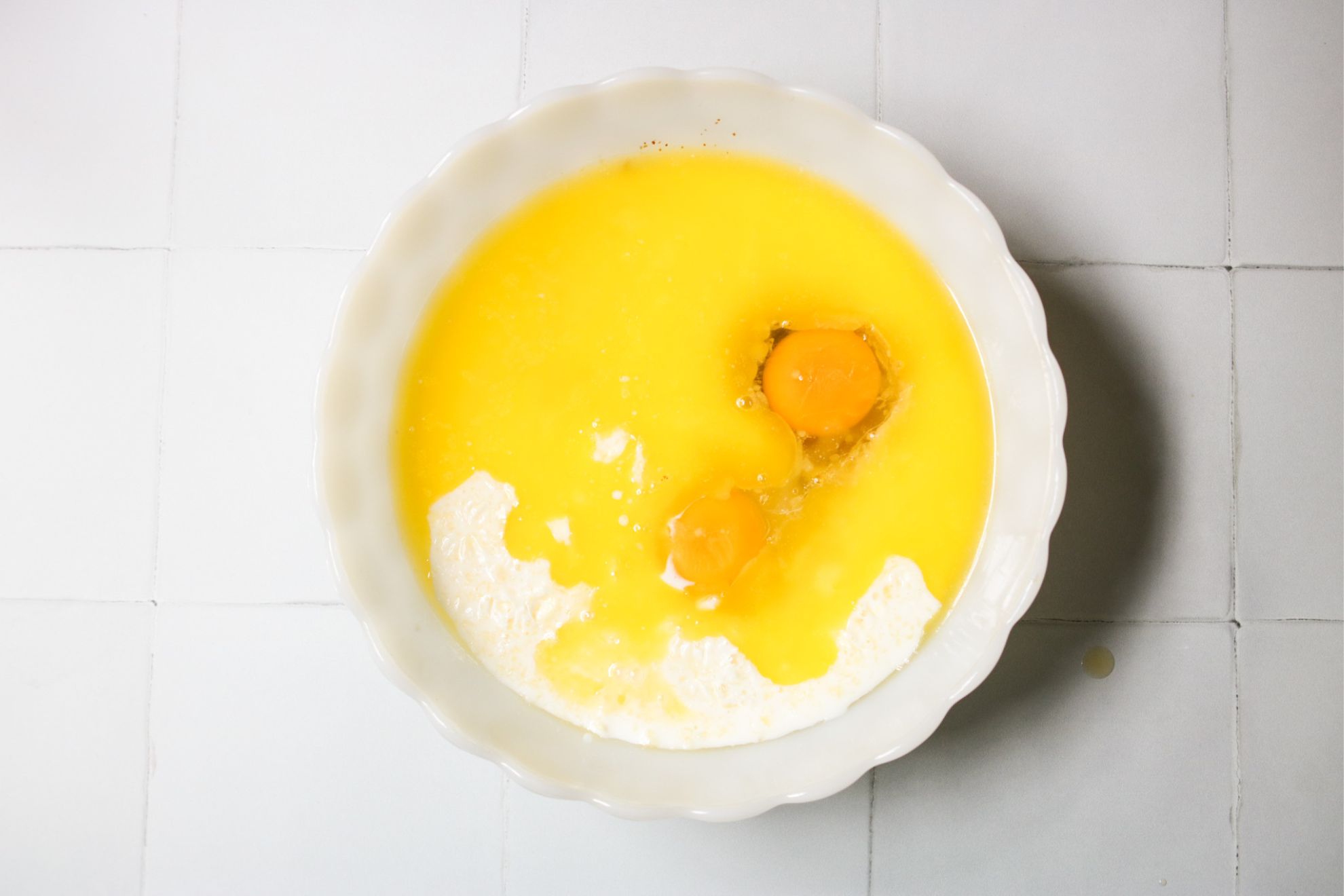 This is an overhead horizontal image of a white scalloped bowl with melted butter, eggs, and cream in it. The bowl sits on a white square tiled counter.
