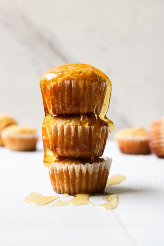 This is a vertical image looking at a stack of three mini muffins from the side. The stack sits on a white surface with more mini muffins are blurred in the background. Honey drizzled on top of the stack with some drips on the white surface around the stack.