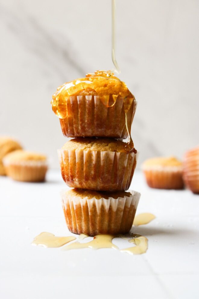 This is a vertical image looking at a stack of three mini muffins from the side. The stack sits on a white surface with more mini muffins are blurred in the background. Honey is falling from the top of the image and drizzling on top of the stack.
