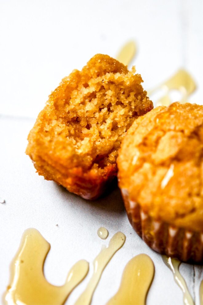 This is an vertical image looking at two mini cornbread muffins from an angled side view. The muffins sit on a white tiled surface. One muffin has a bite taken out of it and is leaning on its side. Both muffins are drizzled with honey, some honey has gotten onto the surface.