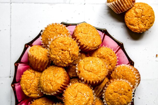 This is an overhead vertical image of a pink glass plate with mini cornbread muffins with white muffin liners and flakey salt. The pink plate sits on a white square tiled surface with more mini muffins to the top left corner of the image.
