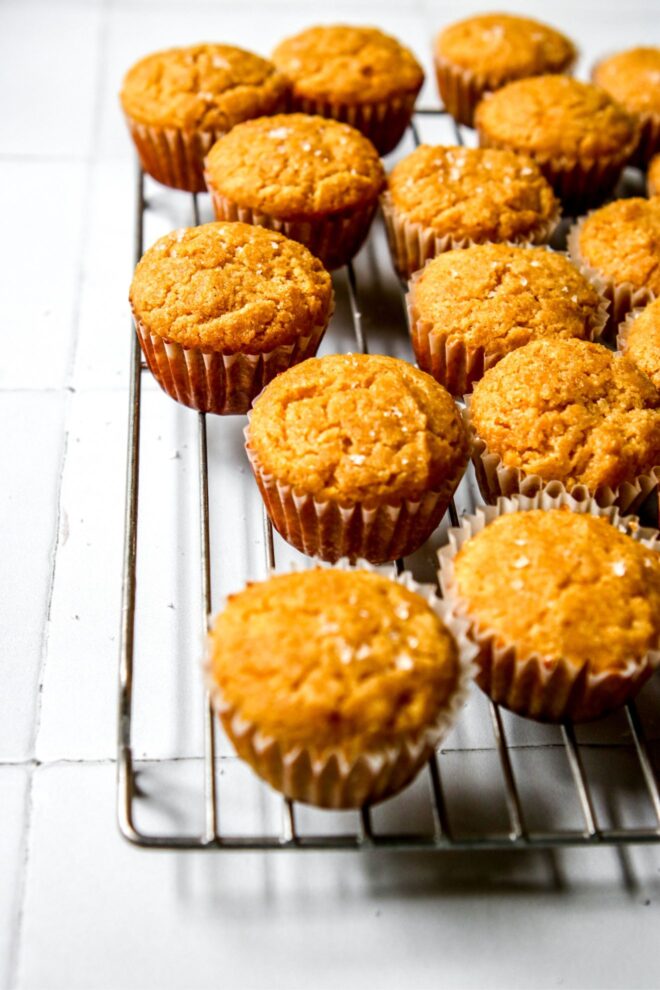 This is a vertical image looking at a silver cooling rack with baked mini cornbread muffins from a side angled view. The mini muffins are topped with flakey salt. The cooling rack sits on a white square tiled surface.