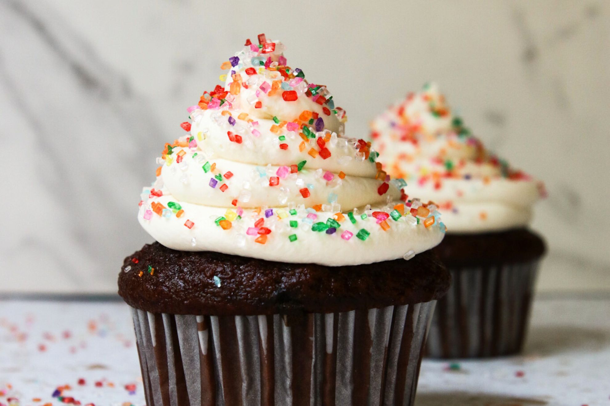 This is a horizontal image looking at a chocolate cupcake from the side. The image focuses on the swirl of vanilla frosting on top of the cupcake. The frosting has rainbow sugar sprinkles on it. Another cupcake is blurred in the background.