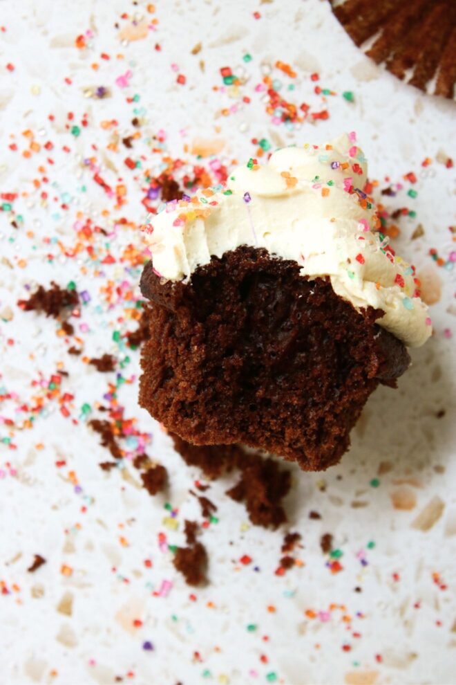 This is an overhead vertical image of a chocolate cupcake with a bite taken out of it laying on its side. The cupcake is topped with vanilla frosting and rainbow sugar sprinkles. More sprinkles are scattered around the cupcake on the terrazzo surface.