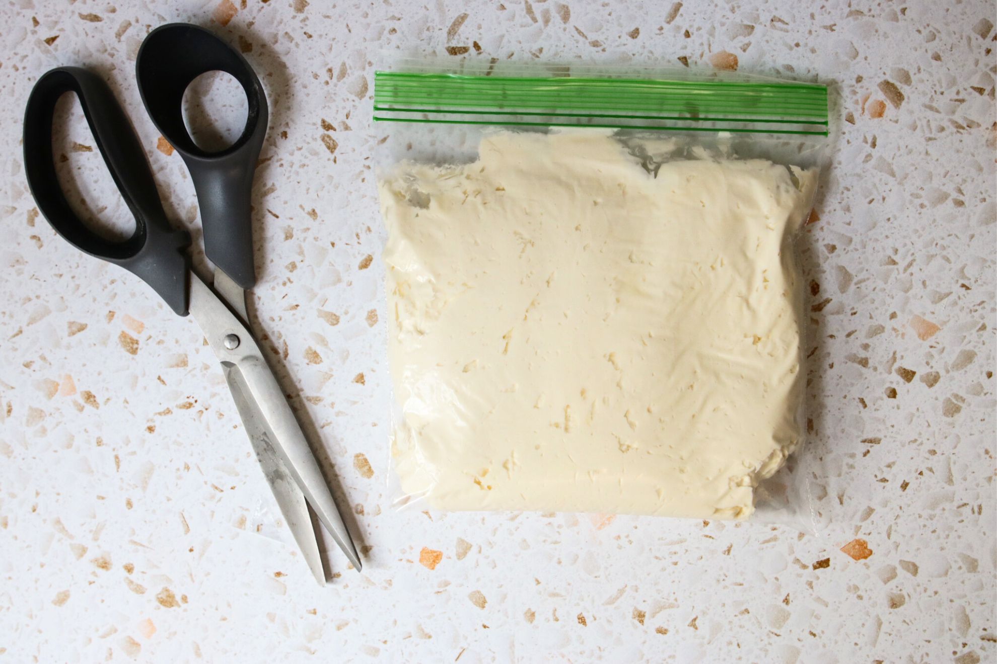 This is an overhead horizontal image looking down on a plastic zip-top bag filled with vanilla frosting on a light terrazzo surface. Scissors are to the left of the baggie.