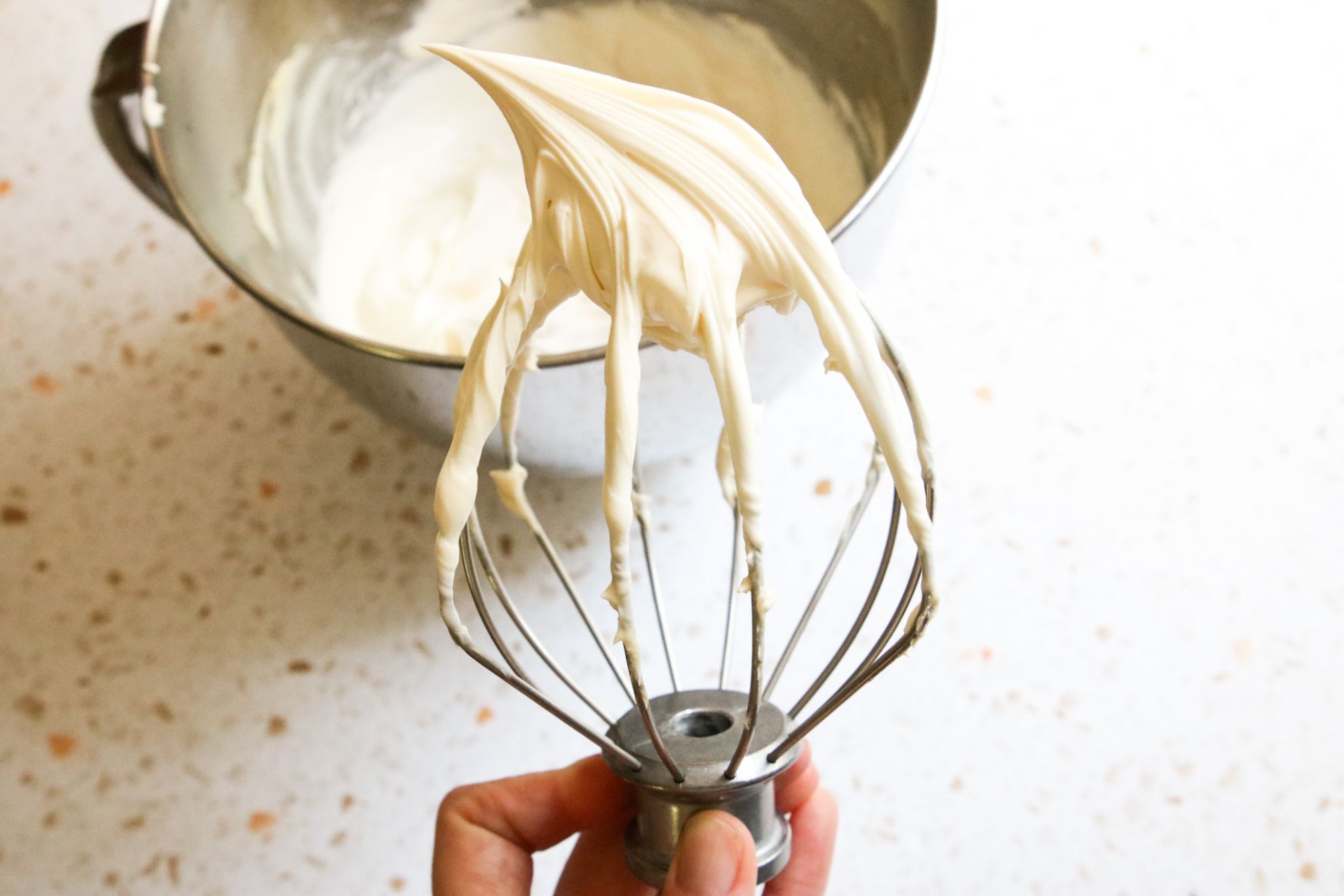 This is a horizontal image of a hand holding the whisk of a standing electric mixer that has been detached. A firm peak of vanilla mascarpone frosting is in the whisk. In the background is a large silver mixing bowl with more frosting on a white terrazzo surface.