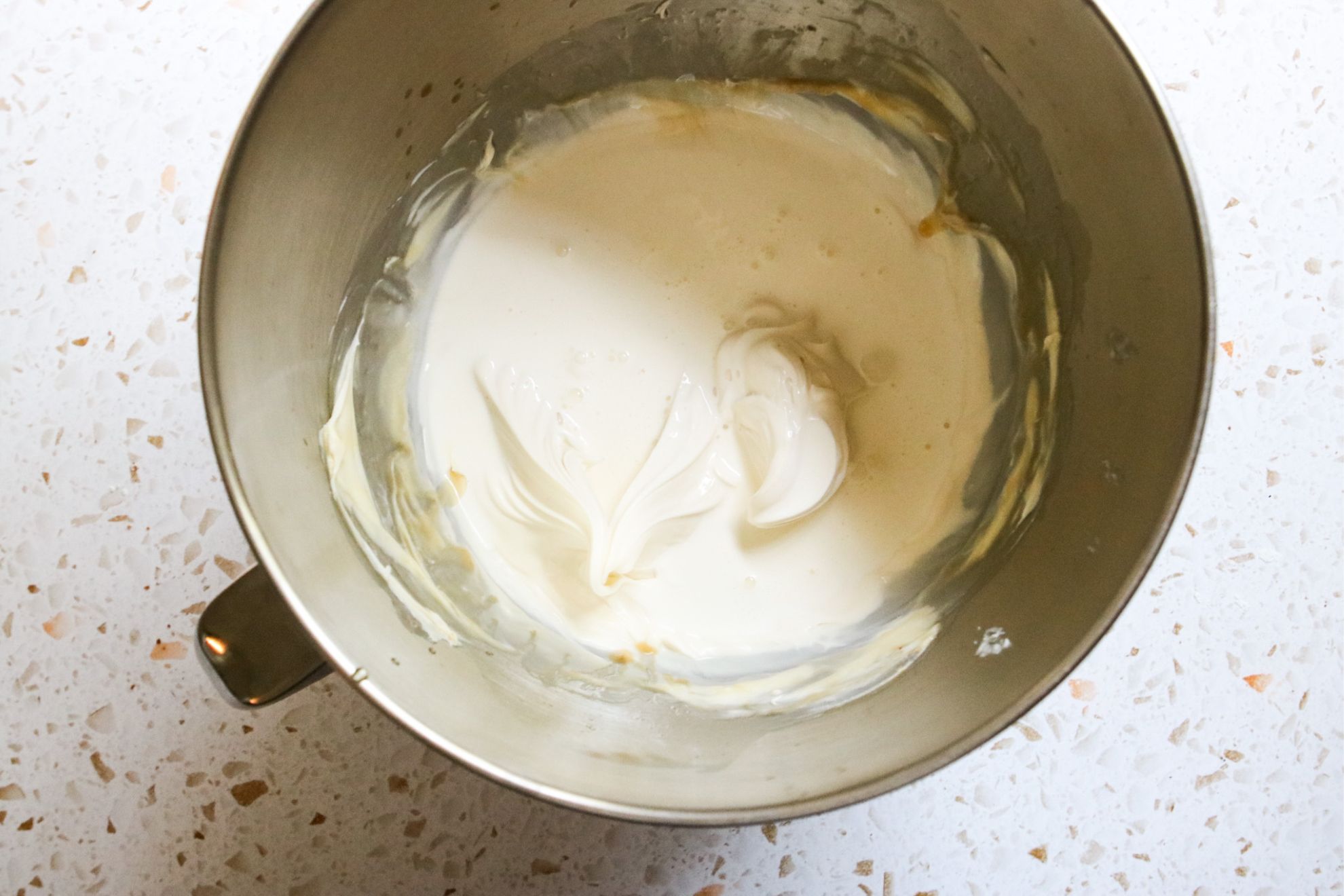 This is an overhead horizontal image of a silver mixing bowl with a vanilla frosting and heavy cream in it. The bowl sits on a light terrazzo surface.