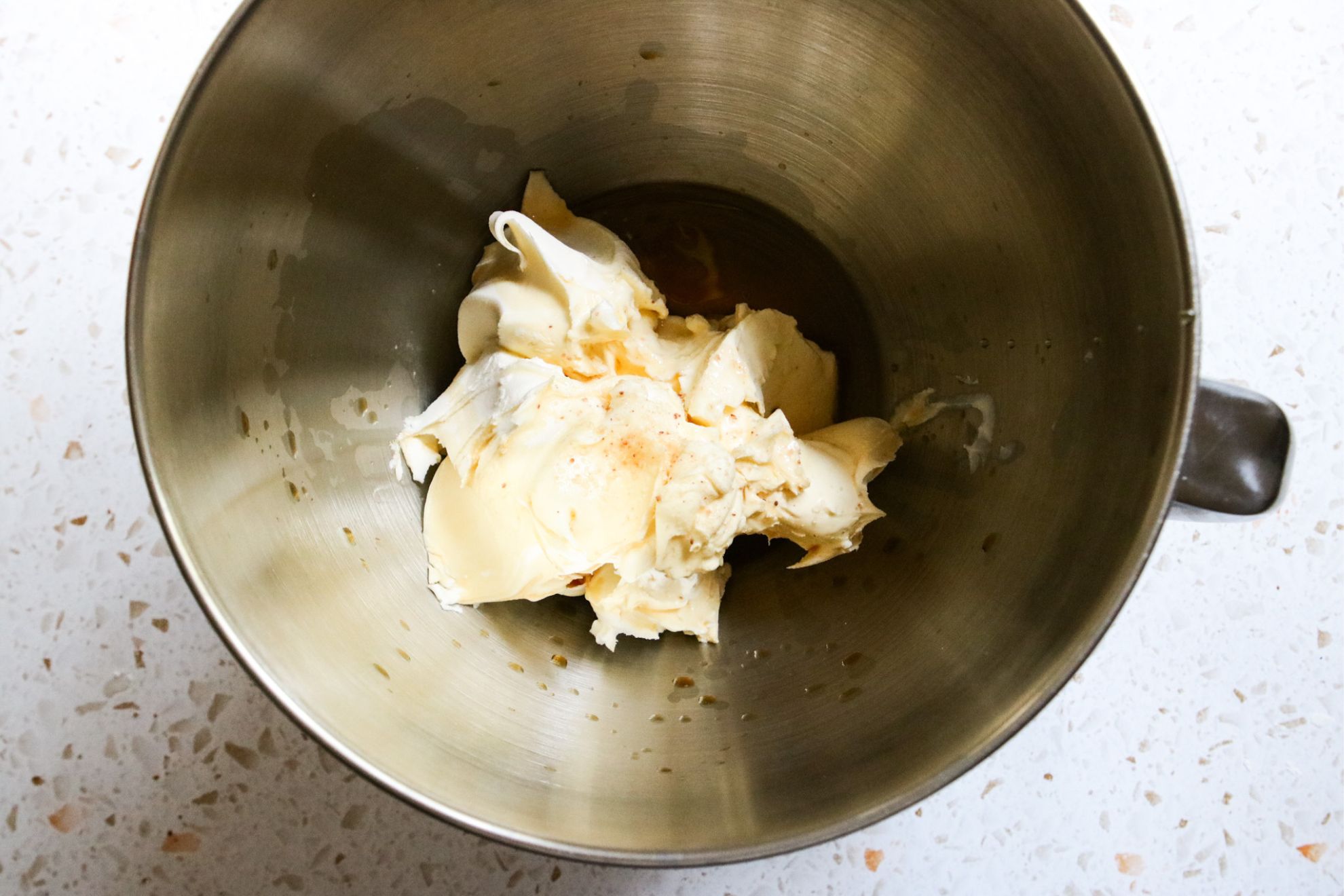 This is an overhead horizontal image of a silver mixing bowl with mascarpone cheese, agave, vanilla and salt in it. The bowl sits on a light terrazzo surface.