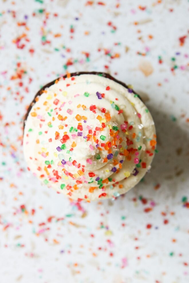 This is an overhead vertical image looking down on vanilla frosting on top of a cupcake. The frosting is topped with rainbow sugar sprinkles with more sprinkles scattered around the cupcake.