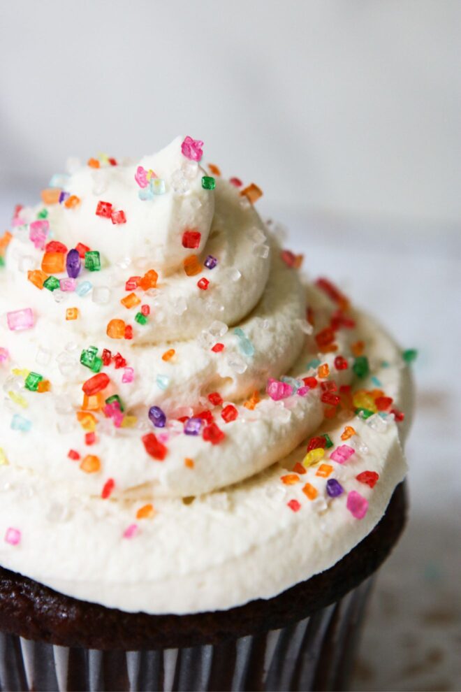 This is a vertical image with a closeup of vanilla frosting on top of the chocolate cupcake. The image focuses on the layers of swirl on top of the cupcake with rainbow sugar sprinkles. The cupcake sits on a white surface with a white background.