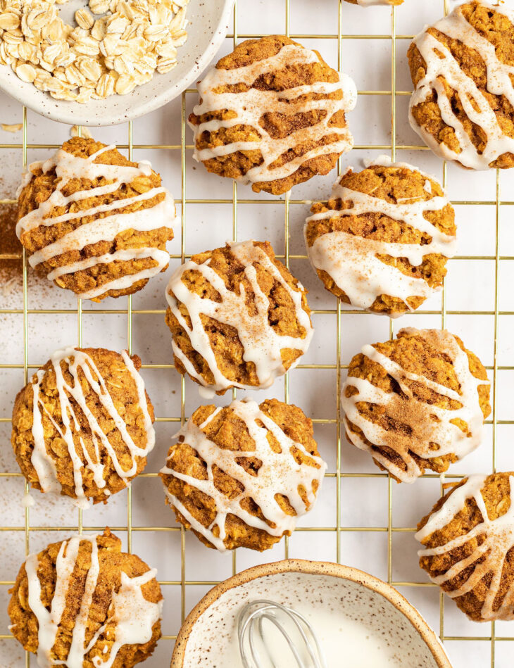 This is an overhead vertical image of pumpkin oat cookies drizzled with a white icing and sprinkled with pumpkin spice. The cookies sit on a gold cooling rack. A small white speckled bowl with oats is to the top left corner of the image and a small white speckled bowl with a brown rim is in the bottom center of the image with white icing and small whisk in it. The cooling rack sits on a white surface.
